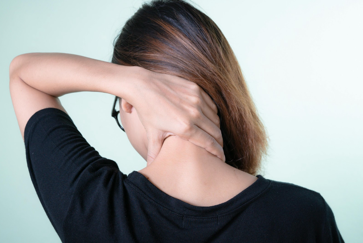 A woman wearing a black shirt holds the back of her neck, appearing to experience discomfort or pain, against a light green background.