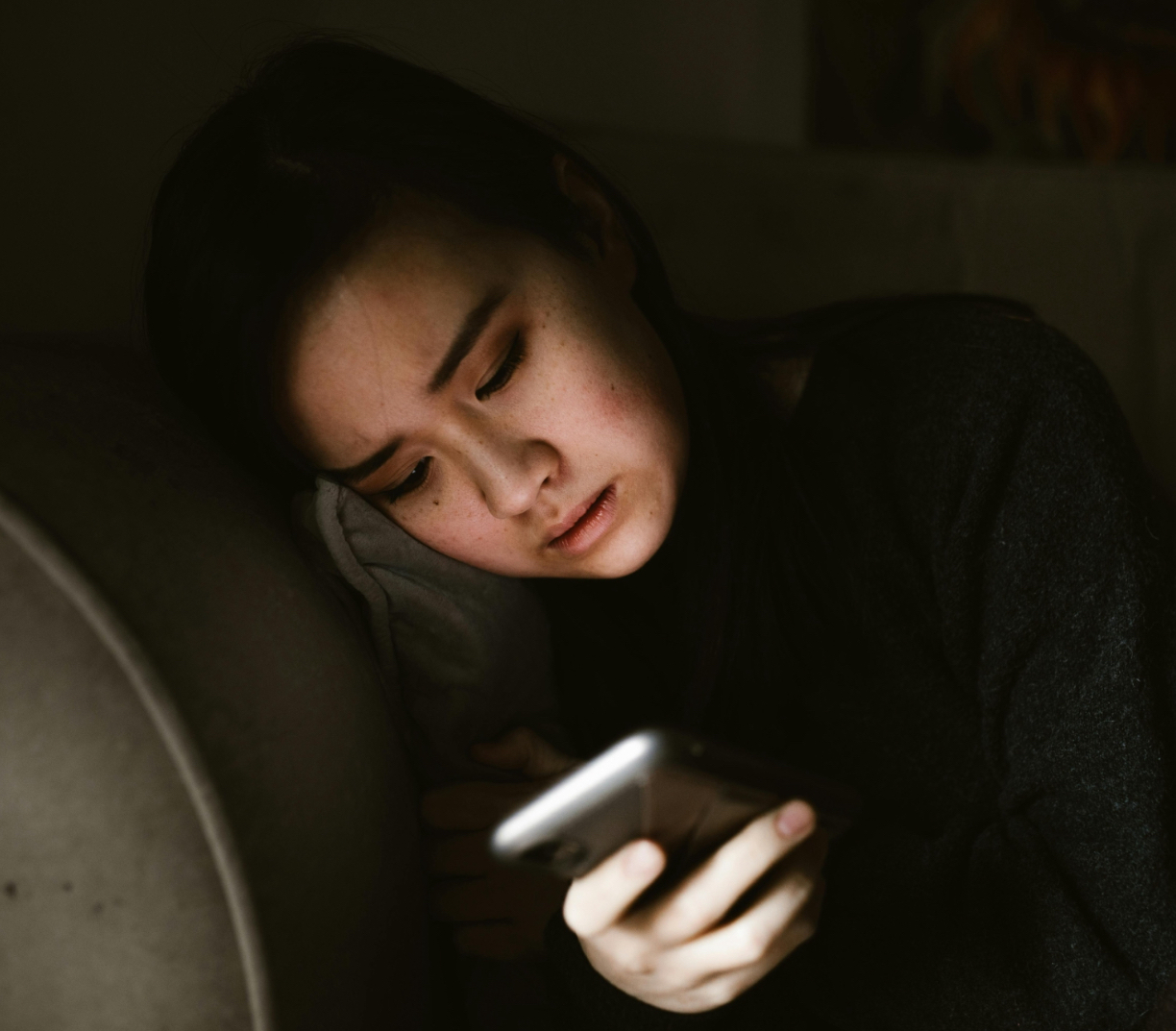 A young woman lying on a couch in a dimly lit room, holding her smartphone close to her face with a strained expression, illustrating the effects of prolonged screen time and poor posture.